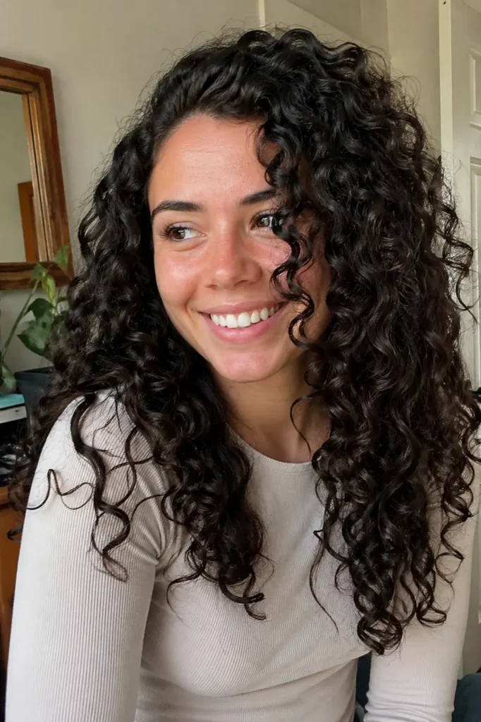 A candid, natural-light portrait of a smiling young woman with dark chocolate–colored, tightly defined curly hair. She’s wearing a light-colored shirt, and her expression is relaxed and genuine, as she looks slightly off to the side.