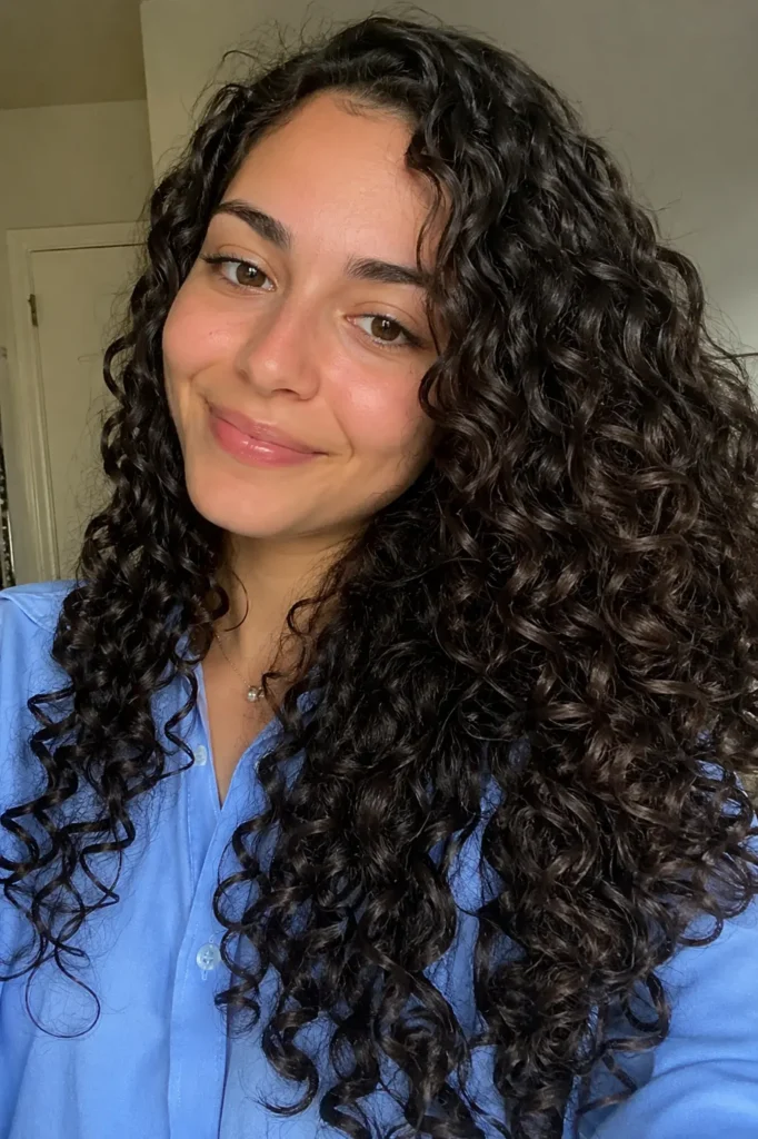 a young woman with defined, glossy spiral curls in a dark chocolate espresso shade. She has a natural smile, minimal makeup, and warm, realistic lighting on her face. She’s wearing a sky-blue shirt,