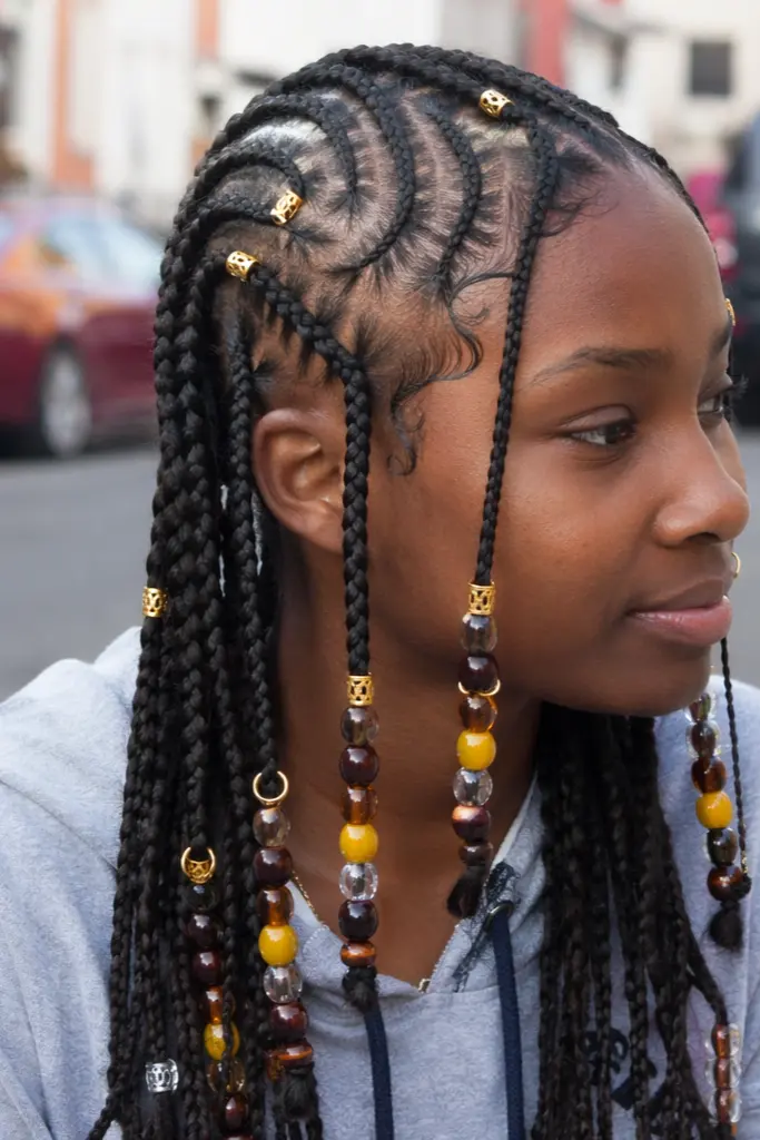 young Black girl with detailed Fulani braids adorned with beads, cuffs, and rings, featuring intricate cornrow patterns on the scalp. She wears a casual hoodie