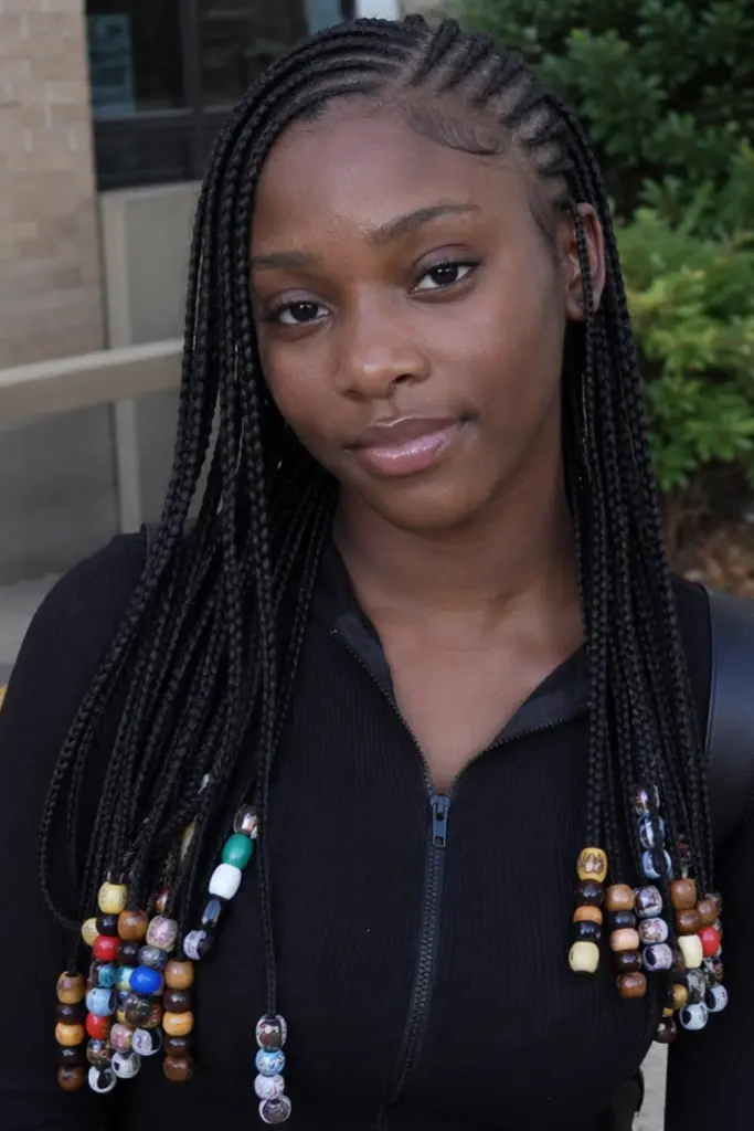 a young woman with Fulani braids styled over a sleek cornrow base, featuring long box braid extensions adorned with colorful and wooden beads at the ends. She wears a simple black outfit and has a natural, relaxed expression