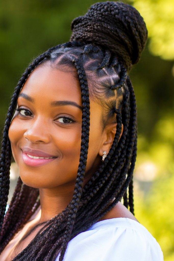 a smiling woman wearing long braided hair styled in a high bun with neat cornrows, standing outdoors in soft natural light with a blurred green background.