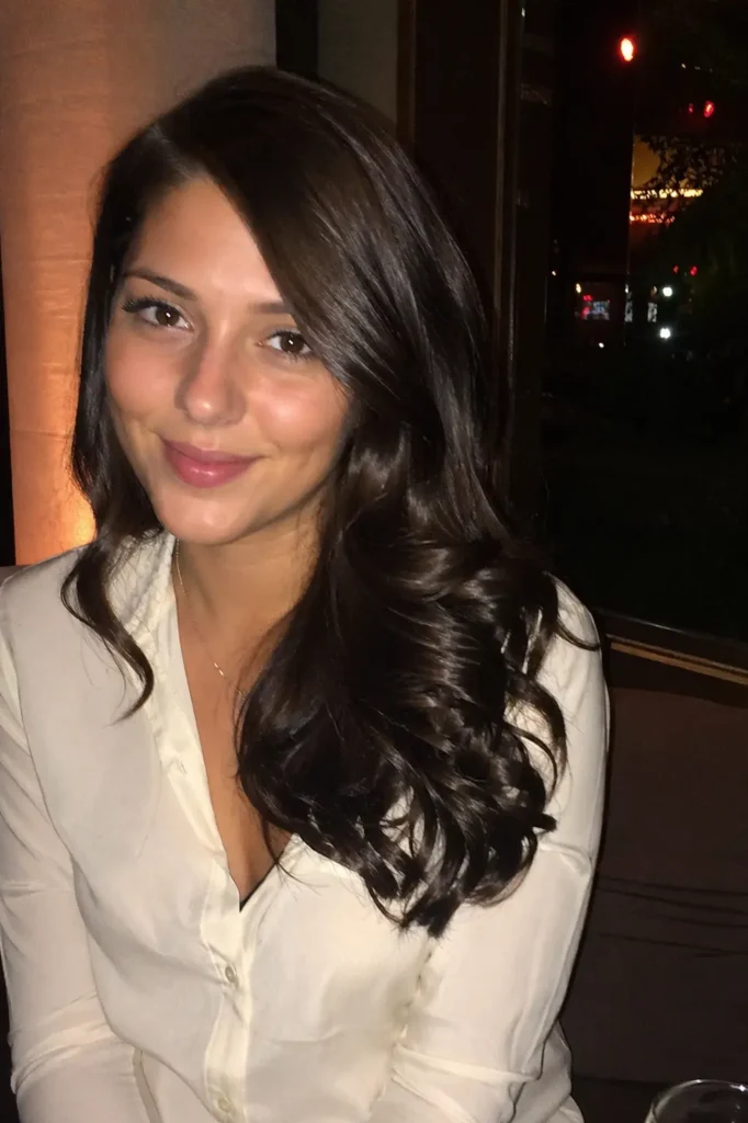 A close-up portrait of a woman with side-parted, glossy dark brown curls, wearing a light-colored blouse. She is seated indoors in a softly lit evening setting, with warm ambient lighting and a blurred background