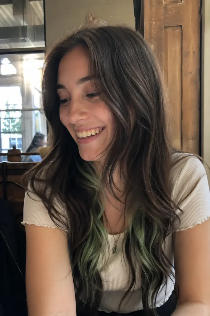 a young woman with long, dark chocolate-brown wavy hair featuring subtle mint-green streaks underneath. She is smiling naturally while looking slightly downward, creating a relaxed, genuine expression. She wears a light-colored shirt and a delicate necklace.