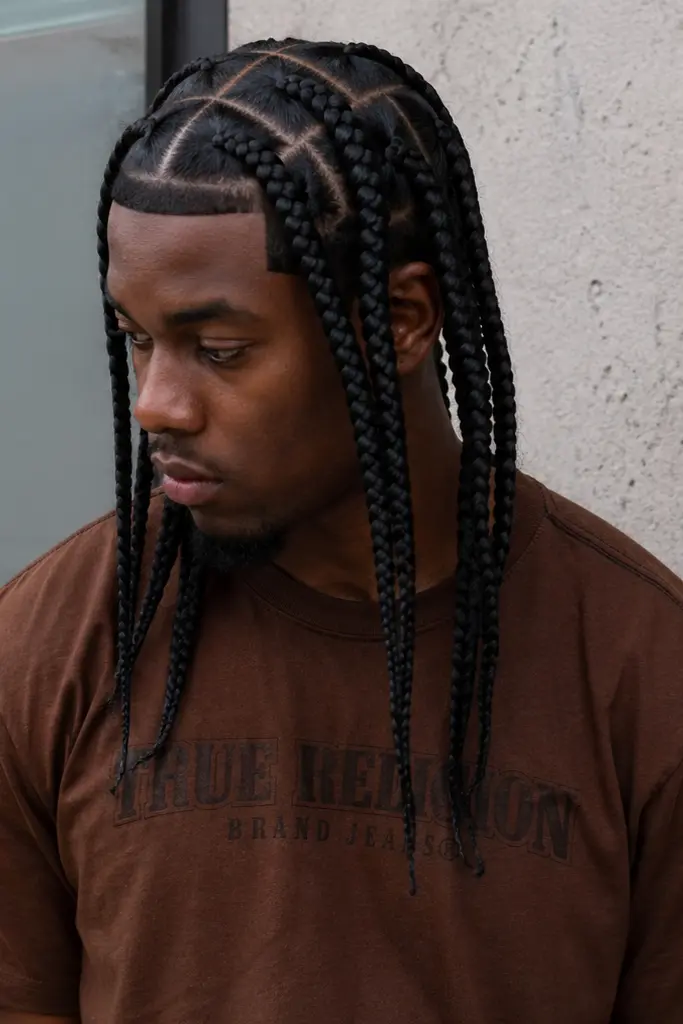 a Black man outdoors wearing a brown shirt, featuring long, neatly parted single braids that hang naturally. His hairstyle is the focal point, with clearly visible scalp sections and a sharp, well-defined hairline.