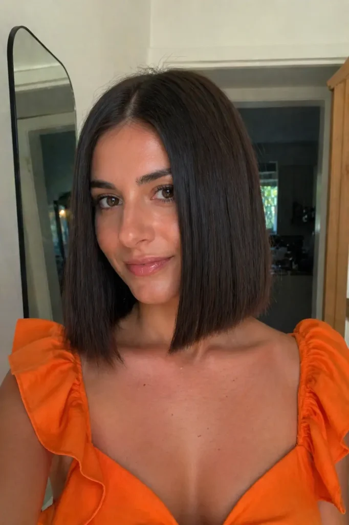 a young woman with a blunt, straight bob haircut and glossy dark chocolate hair, wearing an orange dress, photographed indoors in a modern home setting with soft natural light and minimal background decor.
