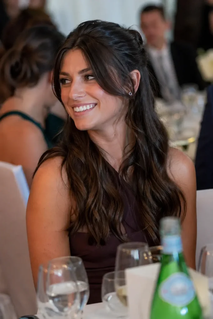 warmly lit event photo of a young woman with long, wavy chocolate-brown hair styled half-up. She is smiling naturally and looking slightly to the side while seated at a table