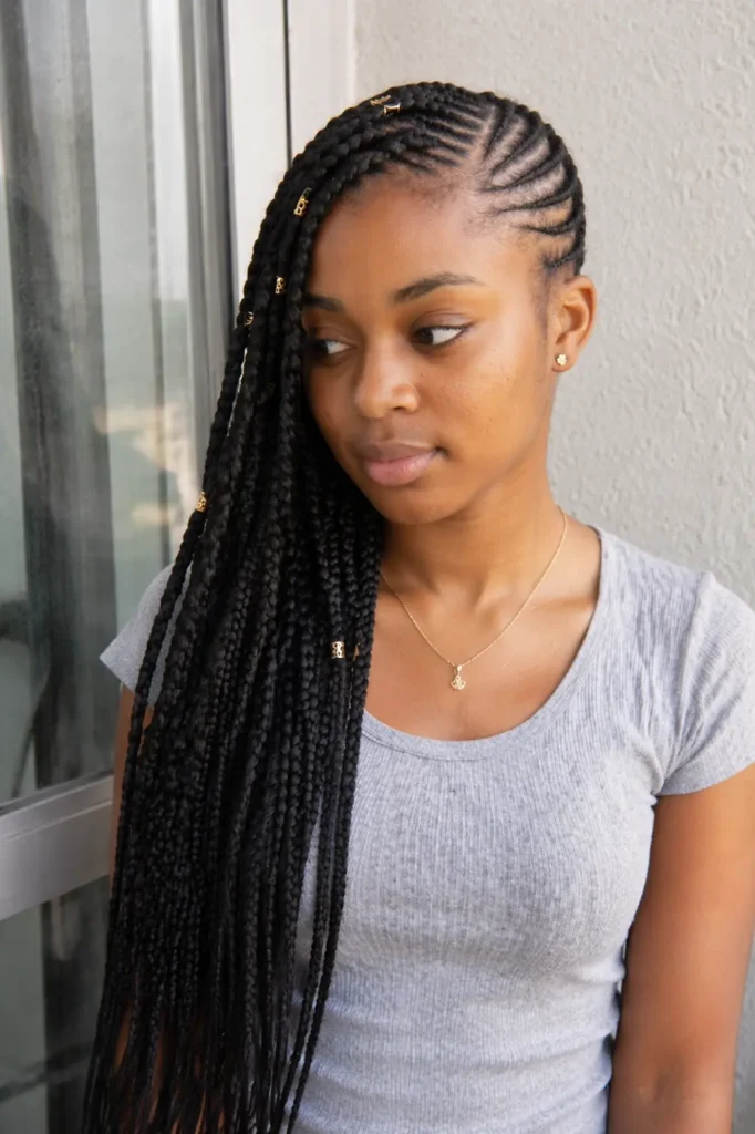 a young woman with side-swept Fulani braids featuring a diagonal cornrow pattern, with long braids draped over one shoulder and accented by subtle gold beads.