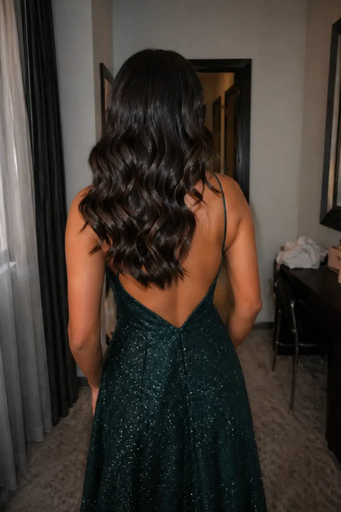 Back view of a real young woman (not a model) with long, soft Hollywood waves in glossy dark brown hair, wearing an elegant sparkly green prom dress with an open back