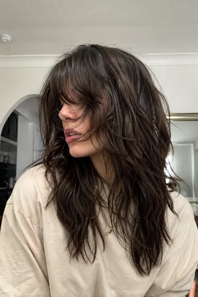 a young woman with dark chocolate brown shag haircut and textured shaggy layers partially covering her face, wearing a light-colored shirt. Soft natural room lighting highlights the realistic hair texture and movement, with a casual side-facing pose and relaxed, everyday atmosphere.