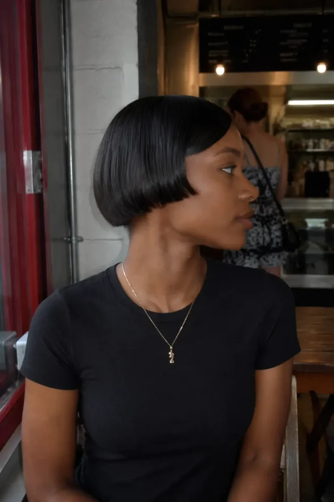 Black woman with sleek blunt micro-bob haircut, straight ear-length hair, wearing a fitted black t-shirt in a casual café setting. Realistic lighting, modern short hairstyle, clean and polished look.