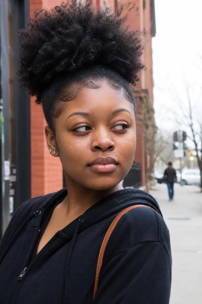 Young Black woman with a high puff afro and defined natural edges, standing on a city street in soft daylight, wearing a black hoodie