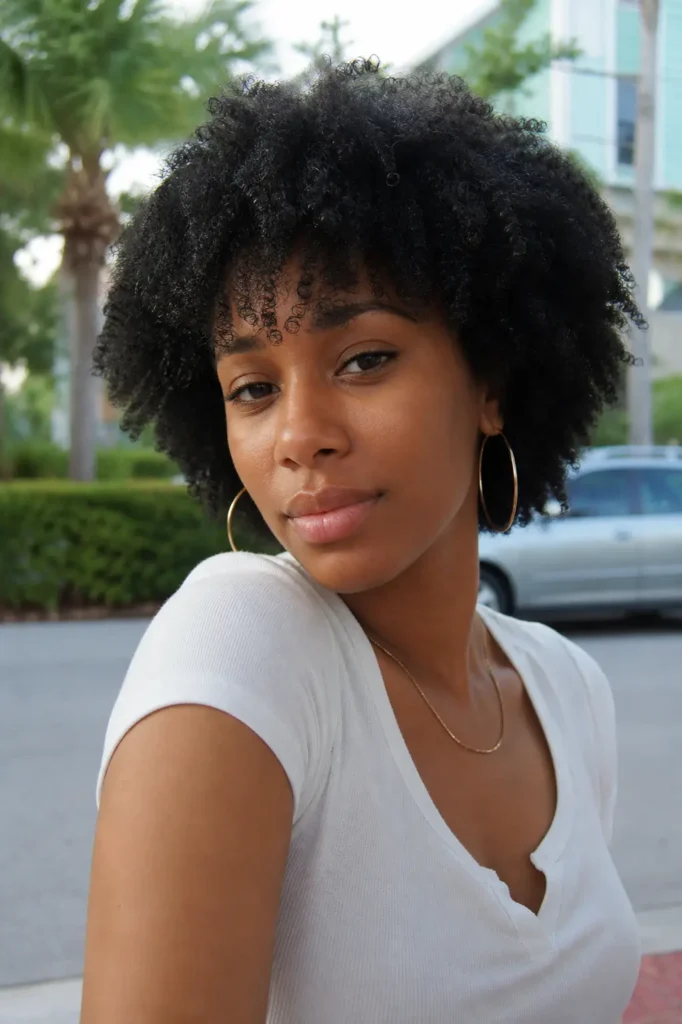 a young Black woman with a natural afro and soft curly bangs framing her forehead. She has smooth, natural skin with minimal makeup and a relaxed, gentle expression