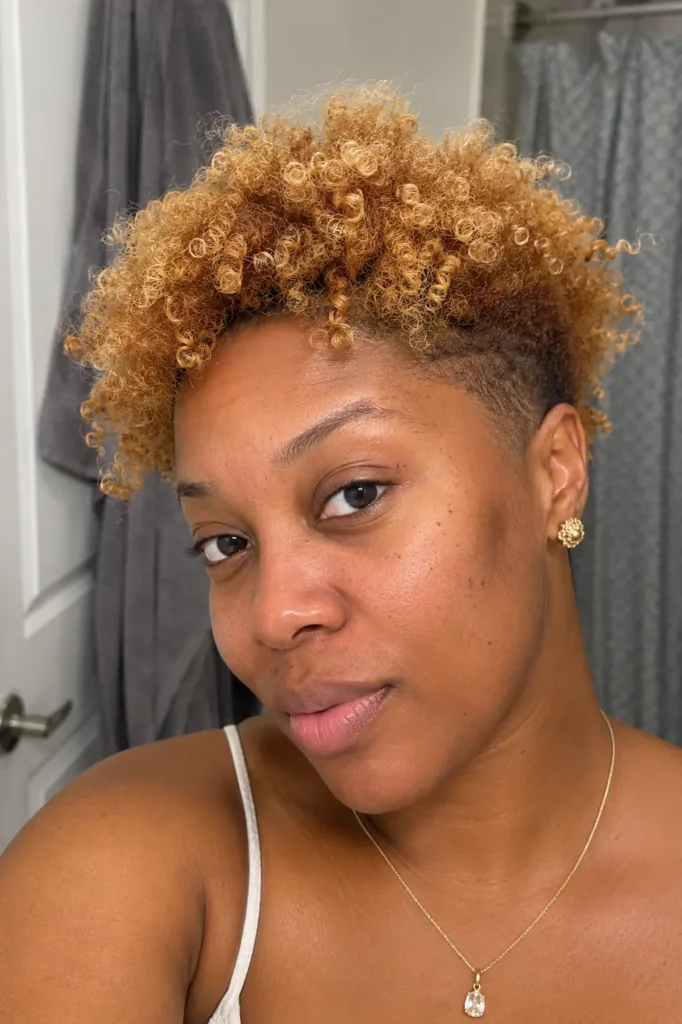 a Black woman aged around 25–35 with a honey blonde curly bald fade hairstyle. The image highlights warm blonde tones and defined curl texture, with authentic facial features, natural skin texture,