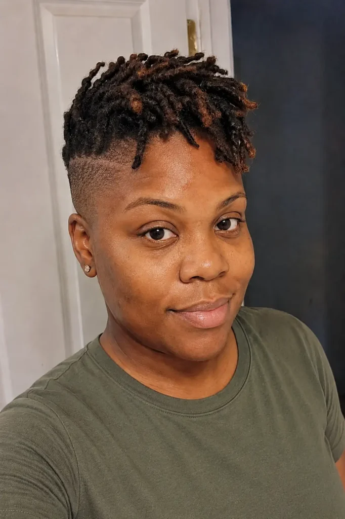 a Black woman aged 25–35 with a short loc top and bald fade hairstyle, featuring visible textured locs and closely faded sides. She has natural facial features, authentic skin texture, a soft everyday expression, and is wearing a modest olive-green shirt