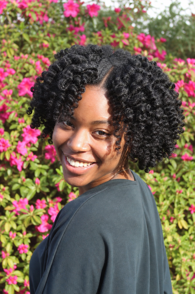 a young Black woman with a bantu knot-out hairstyle, showing defined crimped curls. She smiles candidly in a sunlit garden with vibrant pink flowers in the background,