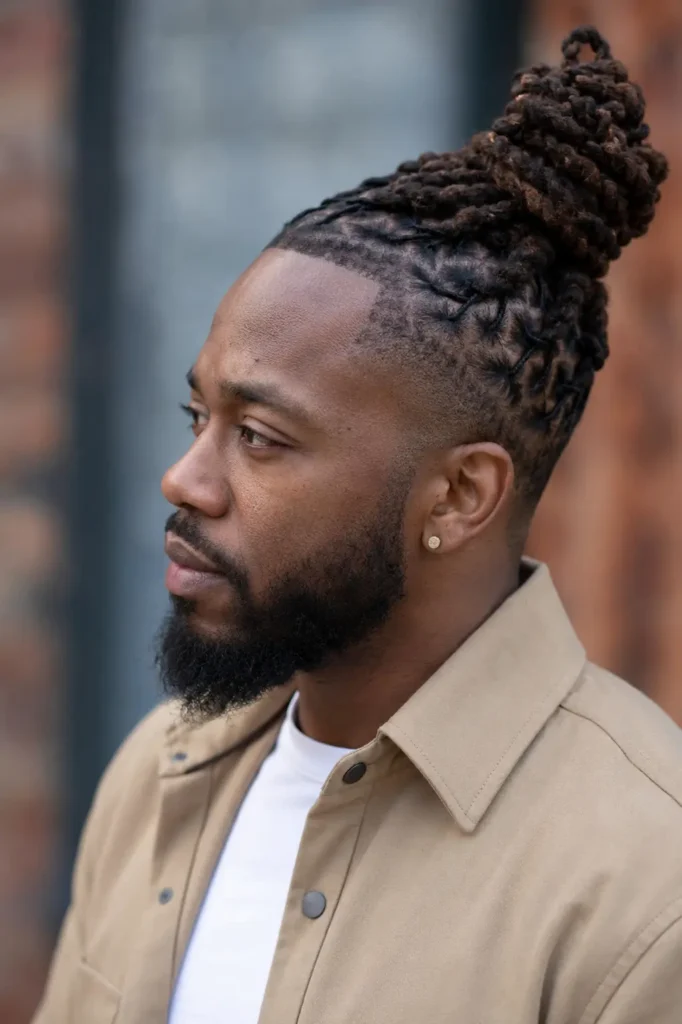 an African American man with a barrel twist loc bun hairstyle gathered high at the back of his head. He is shown in side profile with authentic facial features, natural skin texture, and a neatly groomed beard. Wearing a casual smart beige overshirt over a white shirt, he stands outdoors against a softly blurred urban background