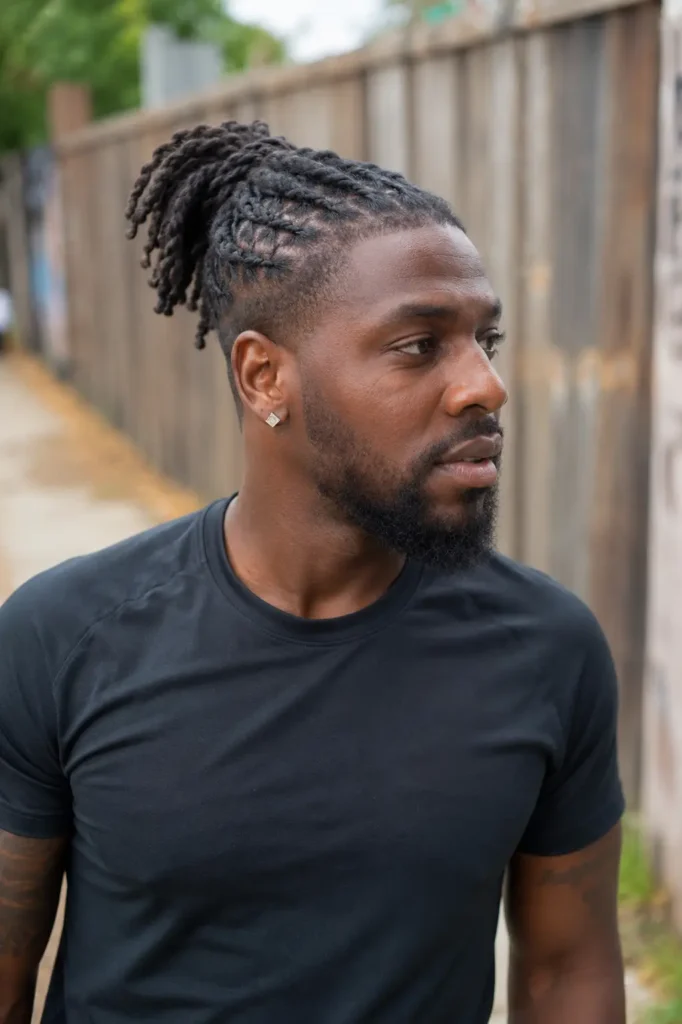 Real Black man with a barrel twist loc ponytail hairstyle standing outdoors in natural light. He has authentic facial features, natural skin texture, a groomed beard, and an athletic build. Wearing a fitted black athletic shirt, he looks to the side with a calm expression