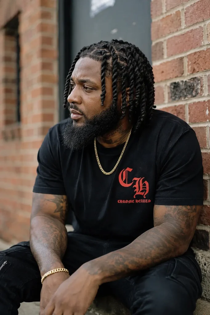 an African American man with a barrel twist loc hairstyle and a full, well-groomed beard. He is seated against a brick wall, wearing a black casual T-shirt and gold chain. His natural skin texture, authentic facial features