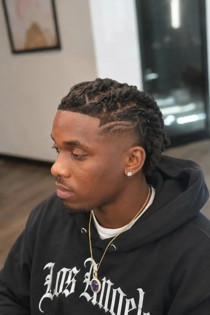 Black man with barrel twist locs styled into neat rows, featuring a shaved line design and a clean fade haircut. He has natural facial features, realistic skin texture, a trimmed beard, and wears a black streetwear hoodie with a pendant necklace