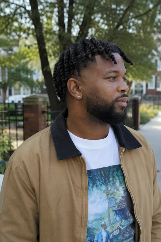 African American man with barrel twist locs and a mid-fade haircut standing outdoors in natural daylight. The portrait highlights the hairstyle's neat twists, shaved sides, and natural texture. He has authentic facial features, warm brown skin with visible natural texture, a neatly groomed beard, and wears a casual modern outfit consisting of a tan jacket over a graphic T-shirt