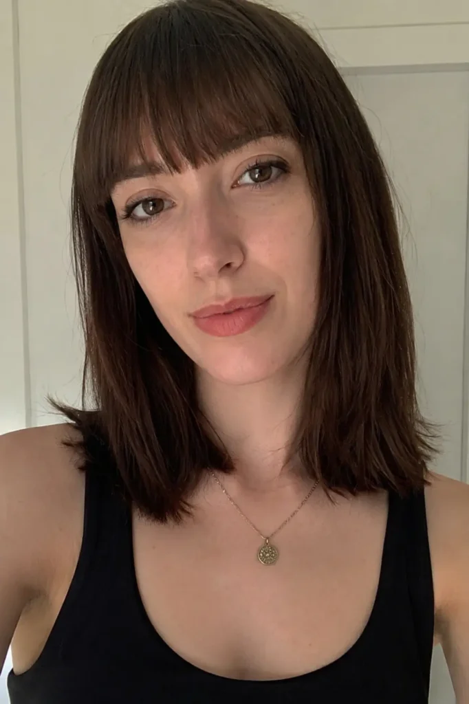young woman with a long face shape and straight medium-length dark brown hair styled with blunt bangs, photographed indoors in soft natural lighting. She wears a black sleeveless top