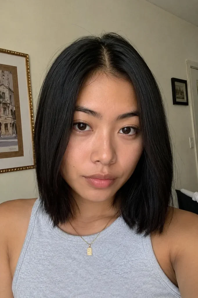 Real girl with a blunt medium-length haircut and middle part, featuring straight shiny black hair and minimal makeup. She wears a neutral gray top