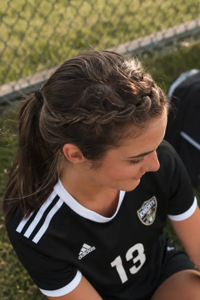 Young woman in a black soccer uniform sitting outdoors on the grass, viewed from above with a side angle, showcasing a medium brown French braid headband hairstyle with visible braid