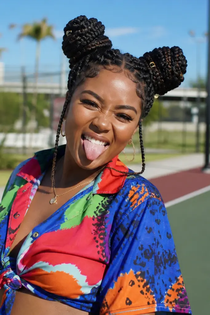 Playful young Black woman with braided space buns, wearing a colorful outfit, posing outdoors in bright sunlight on a park court, smiling with tongue out, with palm trees and a clear blue sky in the background.