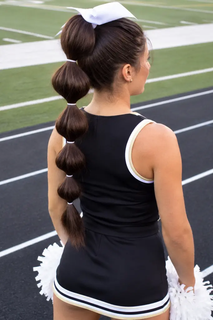 Real teenage cheerleader viewed from slightly above and behind, showing a thick dark brown bubble ponytail with evenly spaced elastics from crown to ends, wearing a black cheerleading uniform and holding white pom-poms on a sports track beside a football field