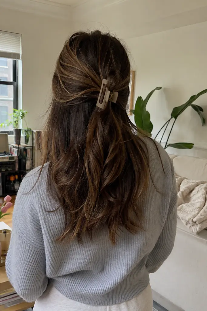 a real woman standing indoors in a cozy apartment living room, wearing a soft casual outfit and a claw clip half-up hairstyle with medium-length textured hair