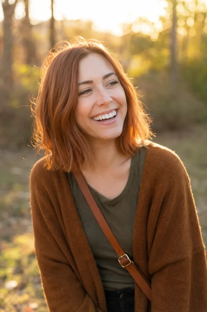 woman in her late 20s with a vibrant copper-toned lob haircut and fine hair glowing in warm golden-hour sunlight, smiling naturally in an outdoor autumn setting. She wears cozy warm-toned clothing and has a genuine happy expression,