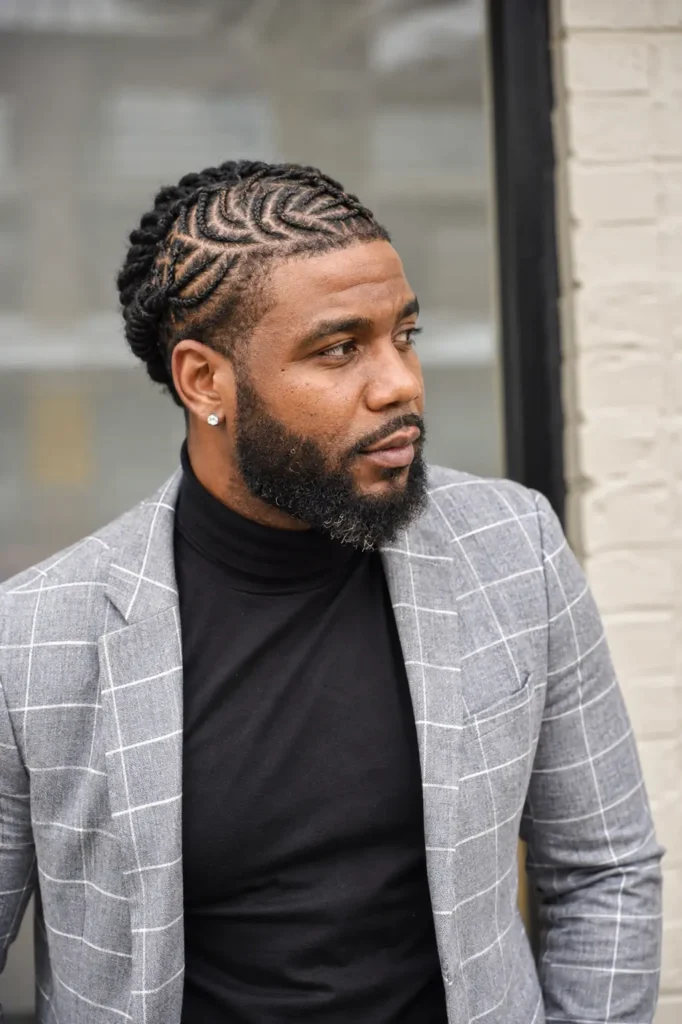 an adult Black man with a crown barrel twist loc hairstyle featuring an intricate circular twist pattern. He has a neatly groomed beard, natural skin texture, and authentic facial features. Wearing a stylish gray plaid blazer over a black turtleneck
