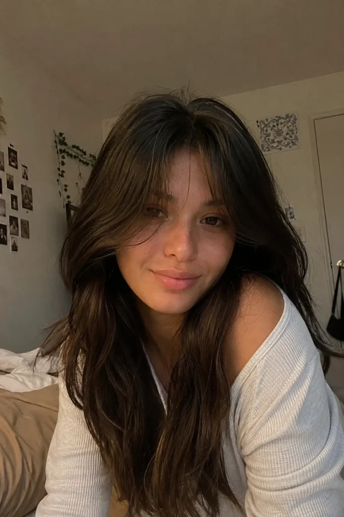 a girl with long layered dark brown hair and curtain bangs parted in the center, posing in cozy indoor lighting with a natural candid expression and realistic hair texture.
