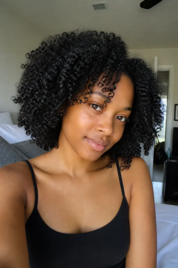 A young Black woman with natural, defined wash-and-go curls poses in a softly lit bedroom, wearing a simple black top and minimal makeup