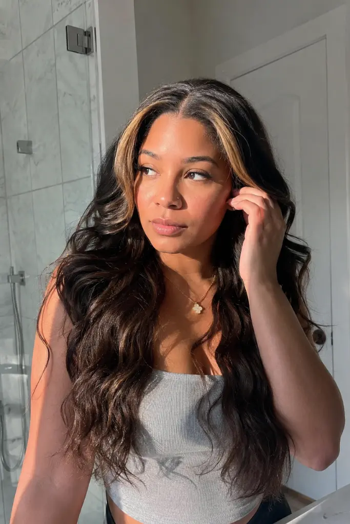 a woman in her early 30s with a medium warm complexion standing in a bright modern bathroom near a mirror. She wears a dimensional dark brown deep wave wig with soft blonde face-framing highlights around the temples and hairline