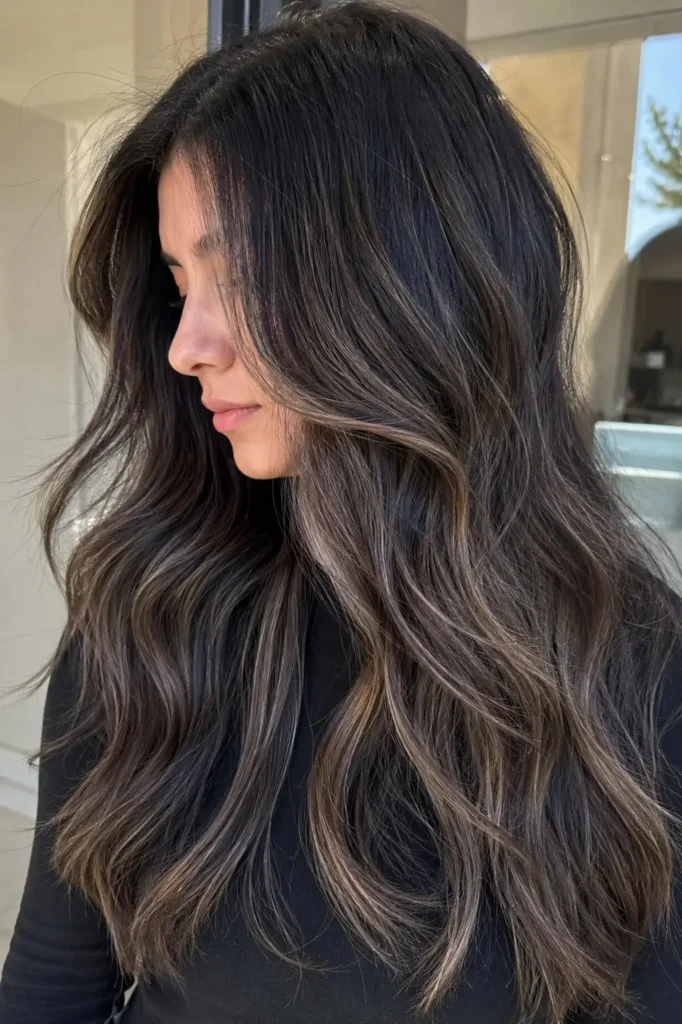 a young woman with long dark hair featuring dimensional cool ash babylights and soft layered waves. Natural lighting highlights the depth, texture, and cool-toned color variation in the hair, which dominates
