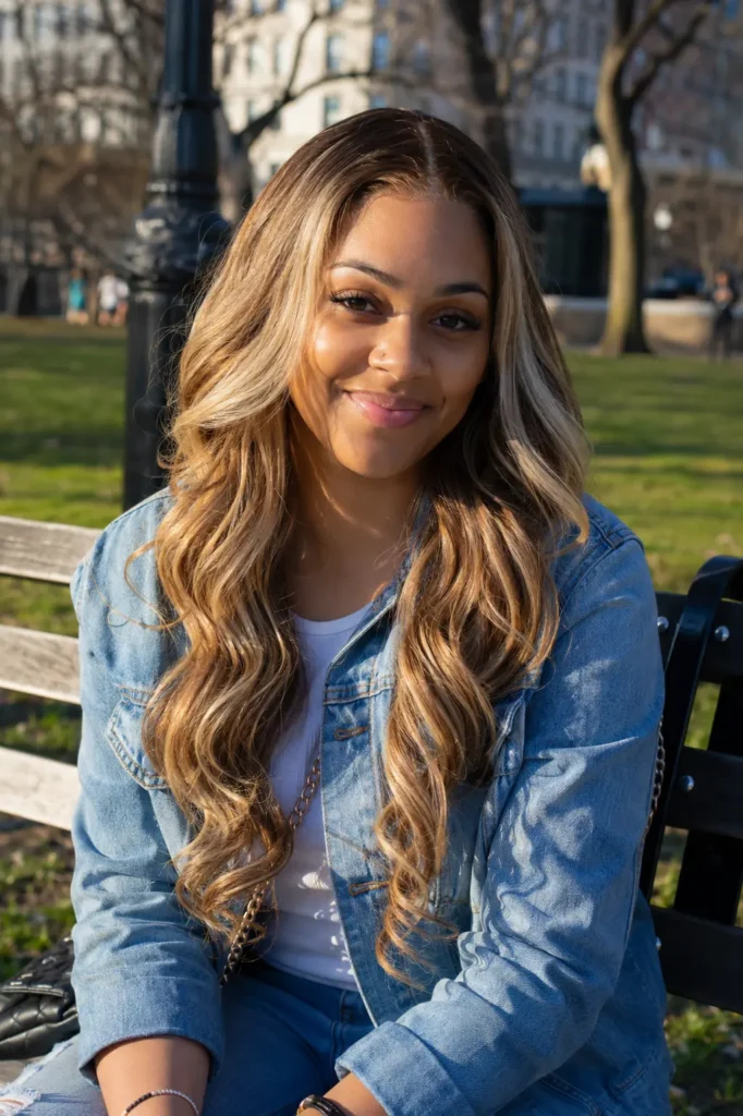 a woman with golden brown skin sitting on a park bench in warm afternoon sunlight, wearing a dirty blonde balayage deep wave wig and a denim jacket