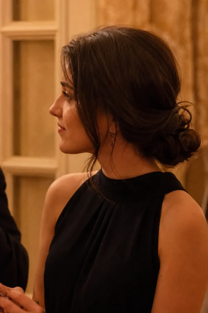 a young woman indoors with an elegant low chignon hairstyle, soft loose face-framing strands, and natural hair texture. She wears a formal sleeveless black outfit