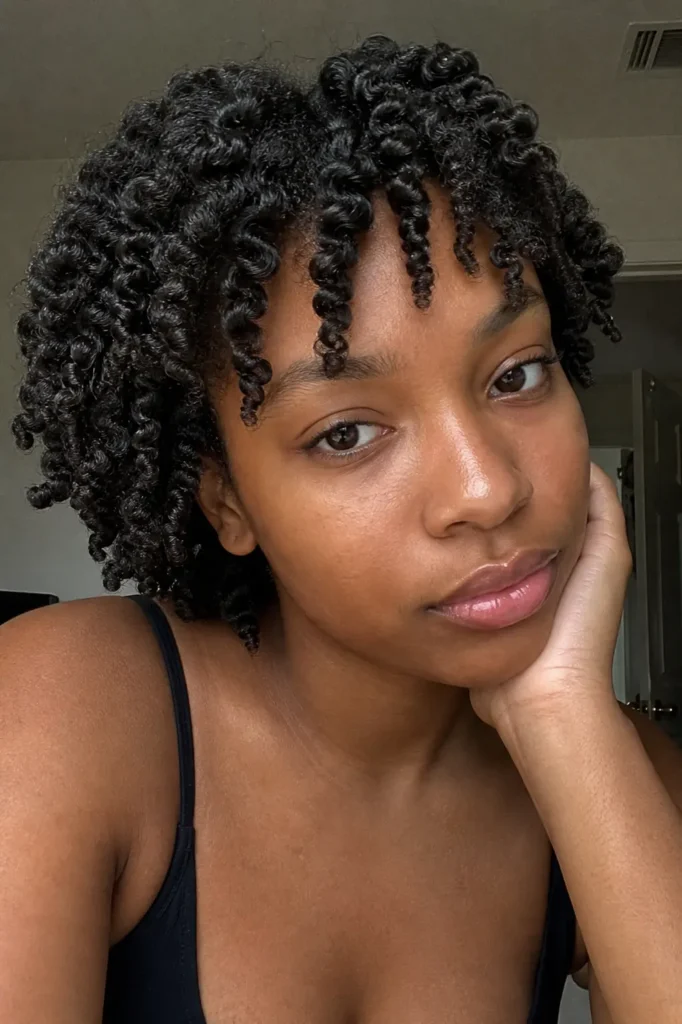 a young Black woman with defined finger-coil curls, resting her cheek on her hand. She has a calm, neutral expression