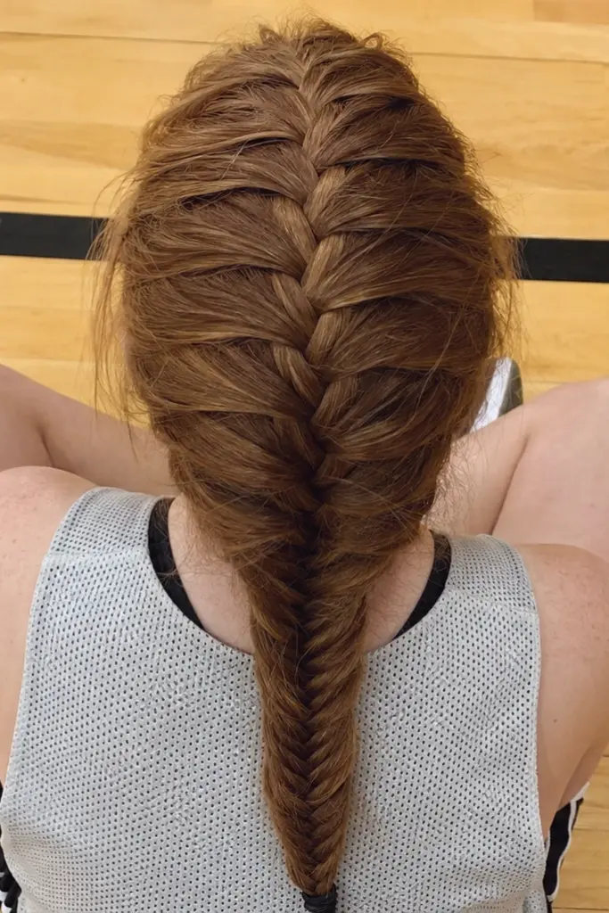 Top-down overhead view of a high school girl with a detailed auburn fishtail braid sitting on a hardwood gym floor in a basketball practice jersey, showcasing a sporty hairstyle.
