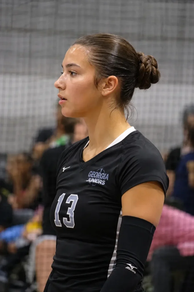 Teen volleyball player with a sleek folded pony bun hairstyle and natural sweat glow, wearing a black jersey on an indoor volleyball court