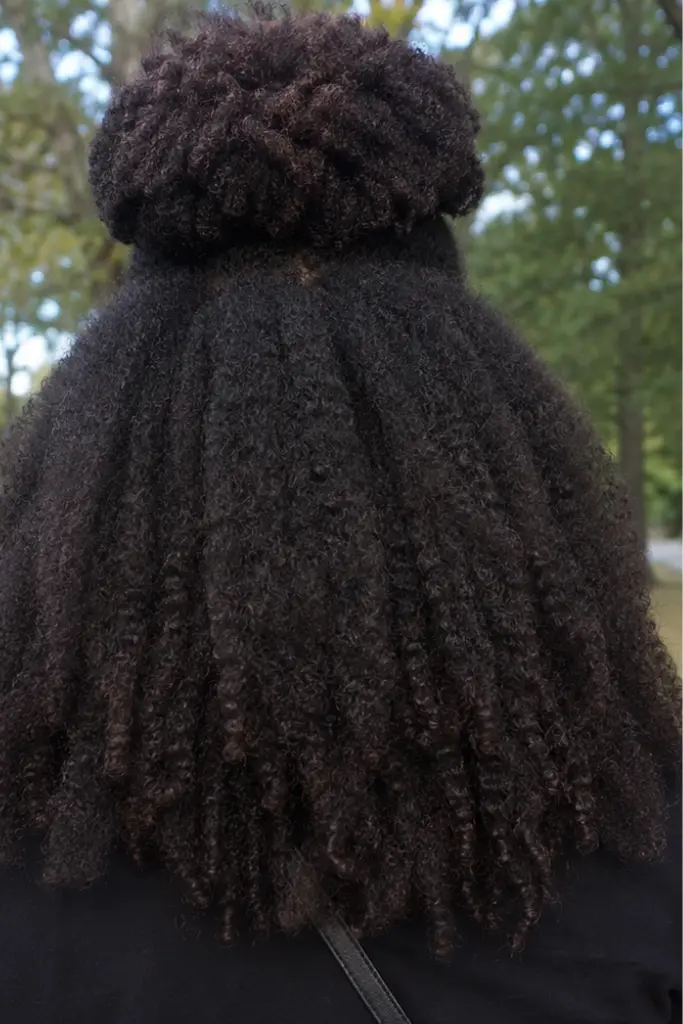 Back view of a real Black woman wearing a natural afro puff half up half down hairstyle in an outdoor park. Her hair features a high voluminous puff on top with long defined coily curls flowing down the back.