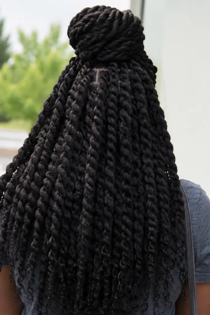 Back view of a woman with jumbo twisted protective hairstyle styled half up in a bun, with large voluminous twists flowing down her back. Outdoor urban setting with natural lighting and a candid, everyday moment feel.