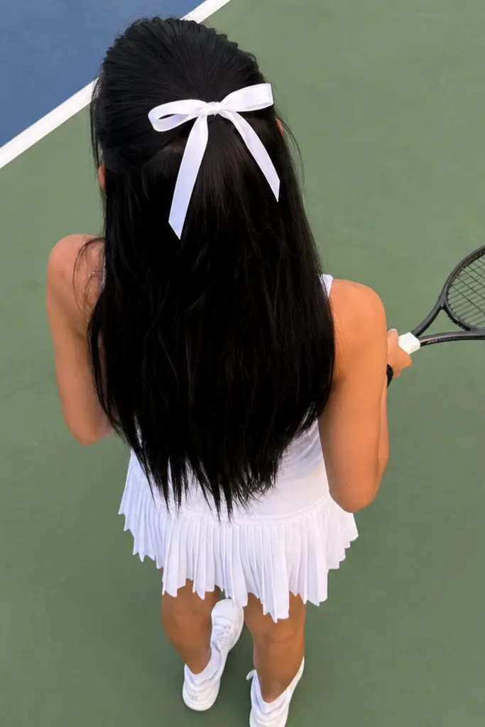 Teen girl with long straight black hair styled in a half-up high ponytail with a white bow, standing on a tennis court in a white tennis outfit, photographed from a top-down back view showing the hairstyle clearly.