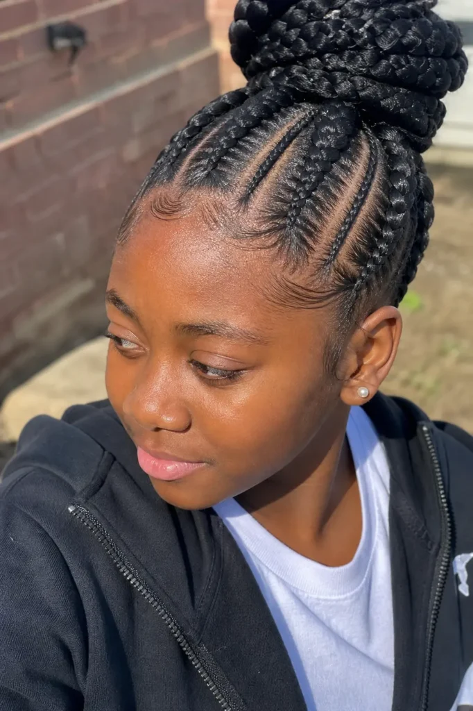 outdoor portrait of a young girl with jumbo feed-in braids styled into a high bun, showing neat scalp parting details. She wears a casual black zip-up hoodie over a white shirt