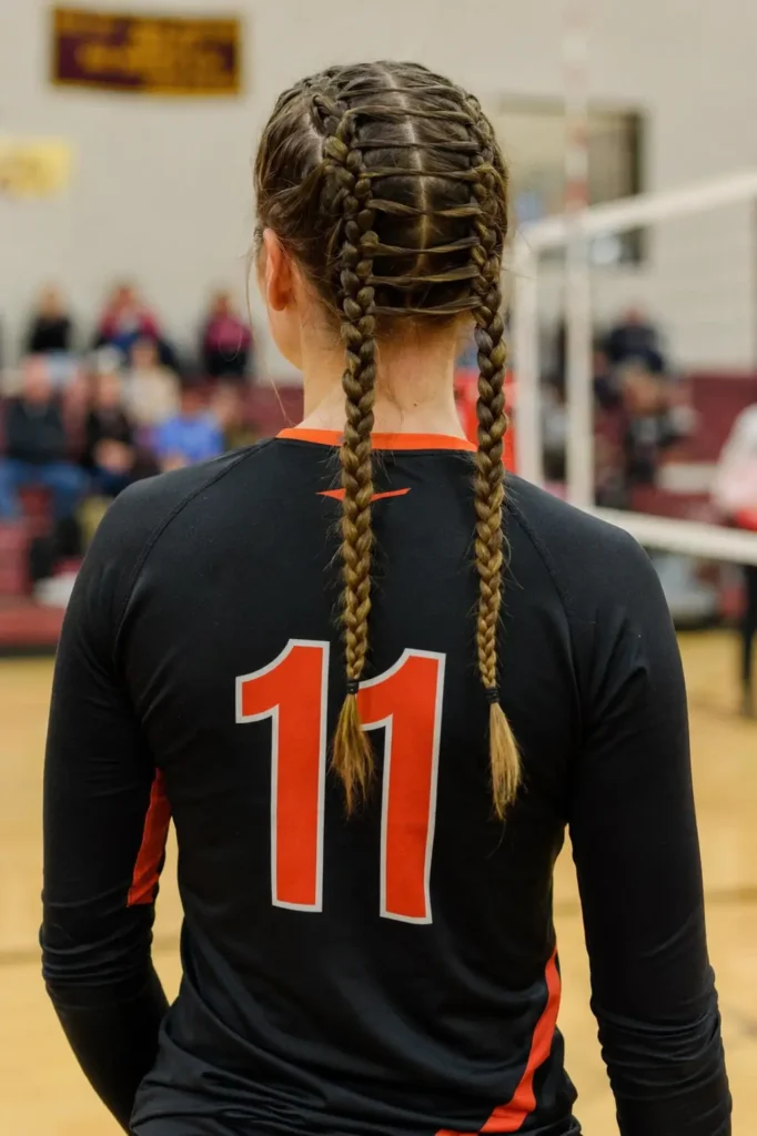 Back view of a young volleyball athlete standing on an indoor court, showcasing an intricate ladder stitch braid hairstyle with parallel braided rows connected by woven ladder-like sections