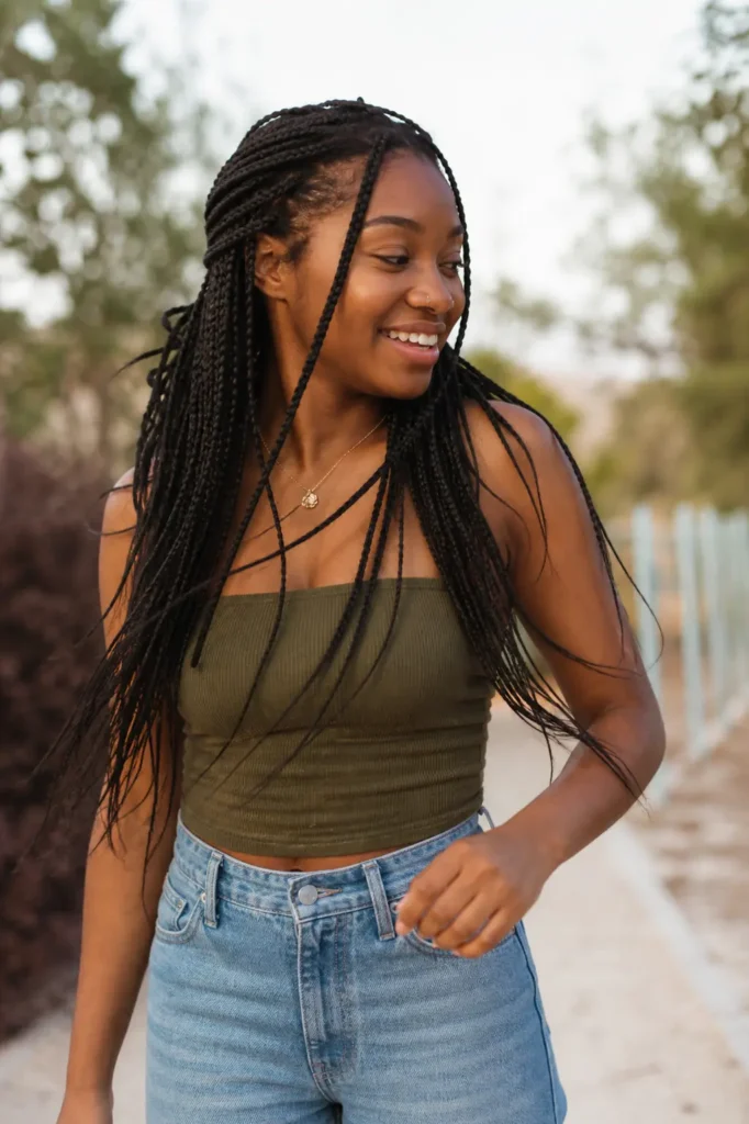 Young Black woman with long layered braids walking outdoors in soft sunlight, wearing a casual green top and jeans, smiling naturally with a candid, relaxed expression against a blurred natural background.