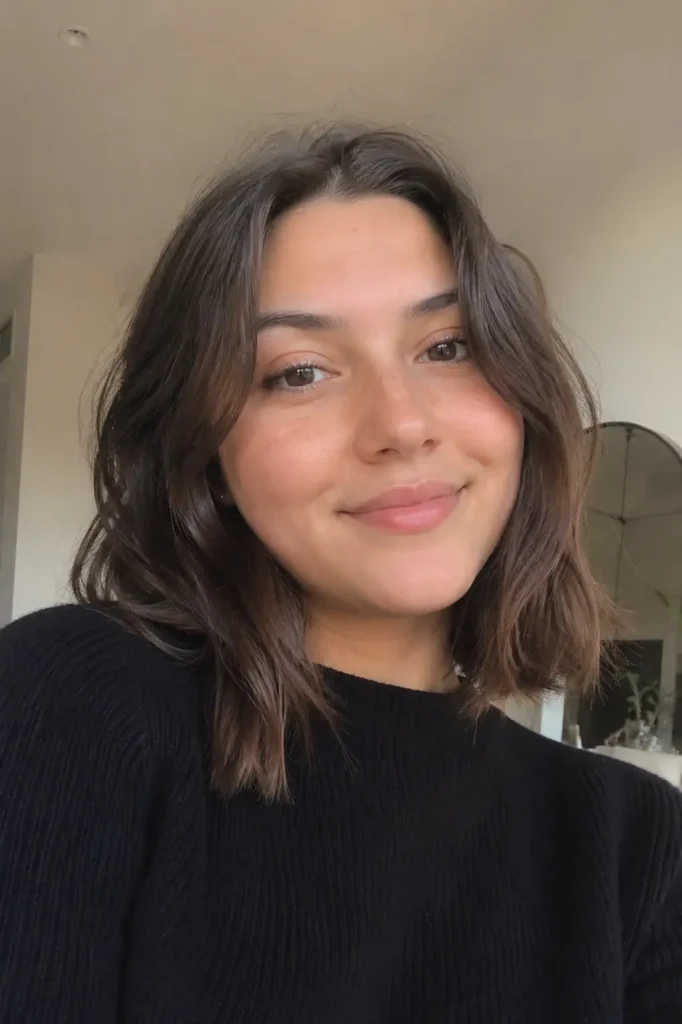 Soft wavy collarbone-length lob hairstyle on a real young woman with a round face shape, photographed indoors in natural daylight. She is wearing a simple black outfit, with realistic hair shine, natural makeup, and a relaxed candid expression in a cozy home setting.
