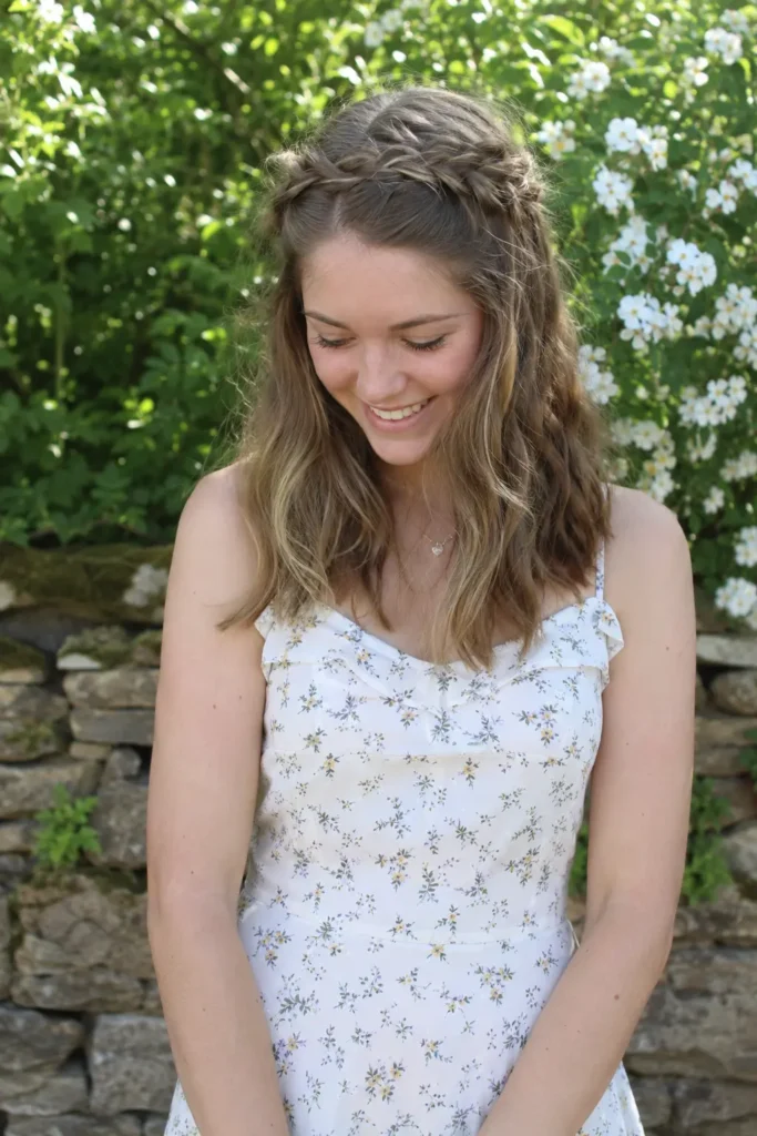 Young woman standing outdoors in a garden, wearing a light floral summer dress, with a loose braided crown hairstyle and medium-length wavy hair,