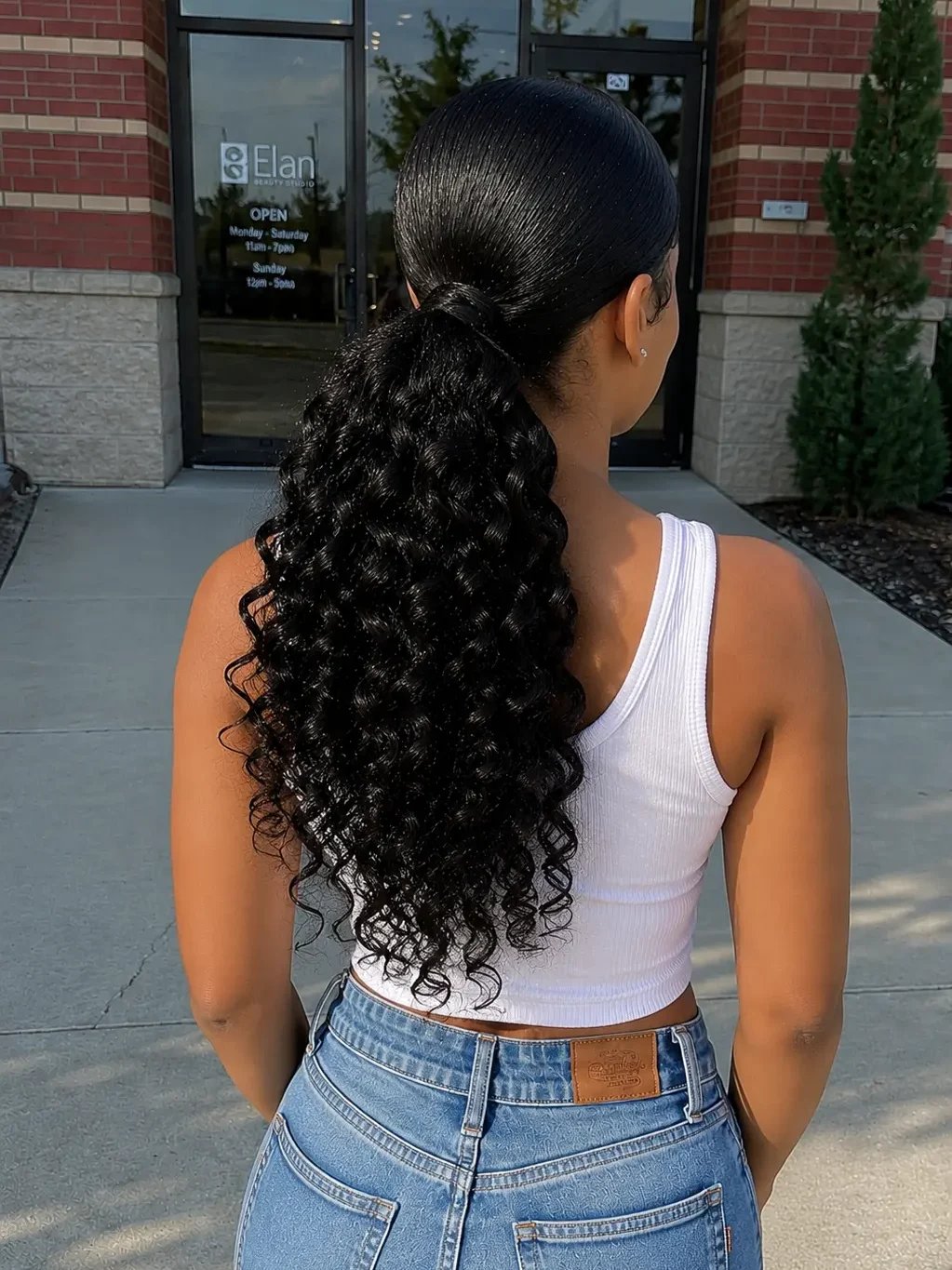 Back view of a real young woman with a low curly quick weave ponytail featuring soft defined curls and movement, wearing a casual white top and jeans